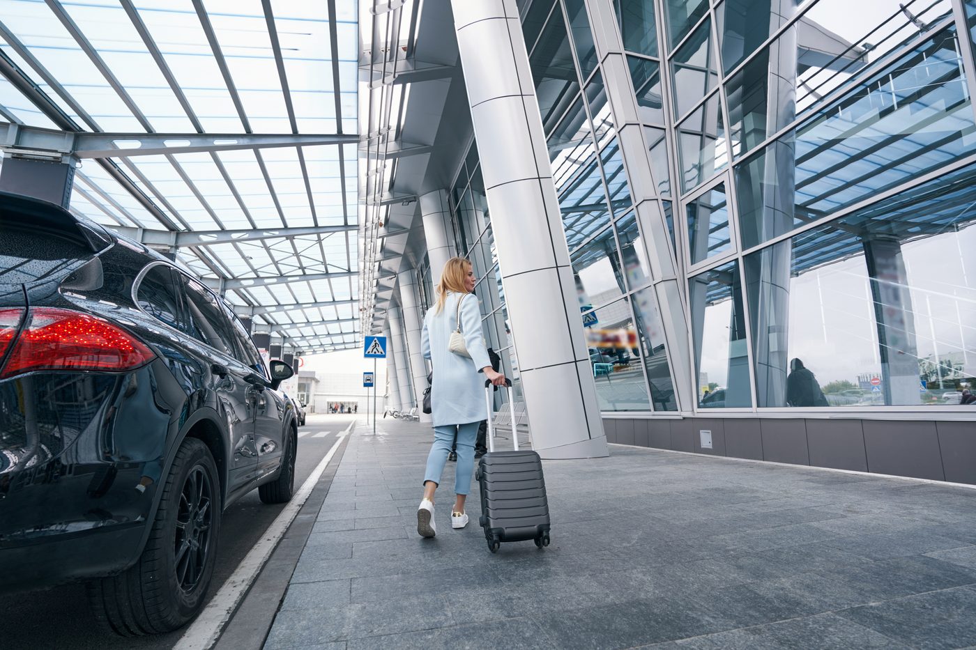 A traveller pulling a suitcase along the curb outside a glass airport terminal