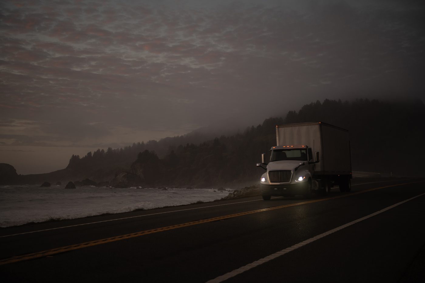 A white box truck with its headlights on driving a coastal highway through evening fog
